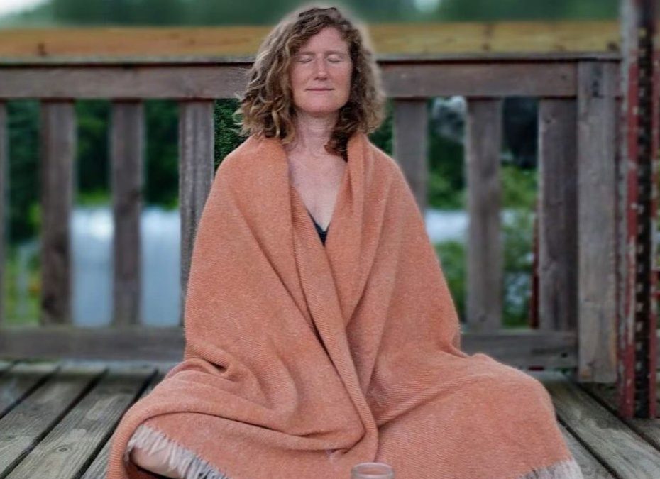 Sarah Belzile sits in meditation on a wooden platform with a peachy-orange shawl draped over her shoulders