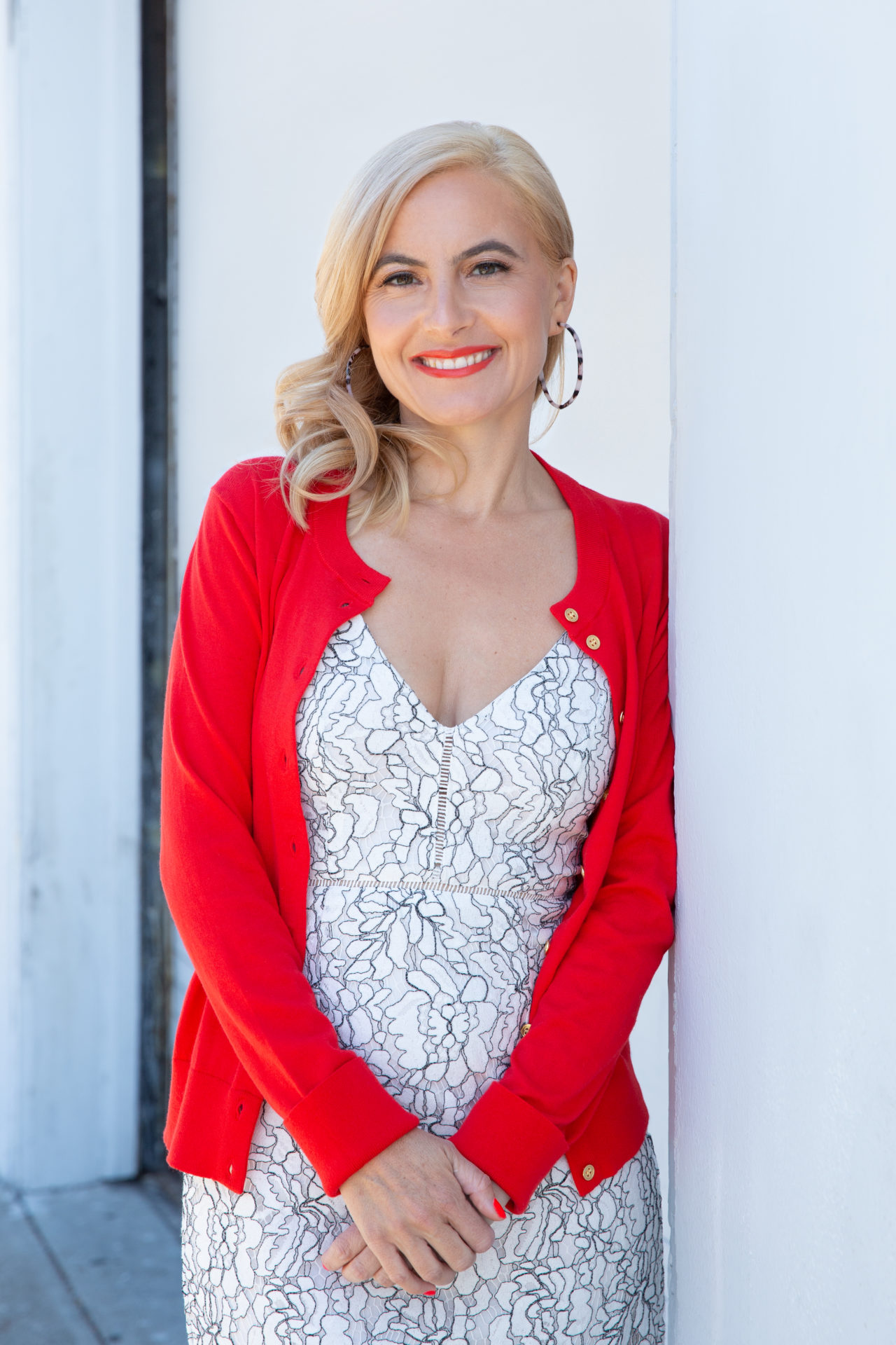 April Hirschman in a white dress with a red sweater leaning against a white wall, smiling.