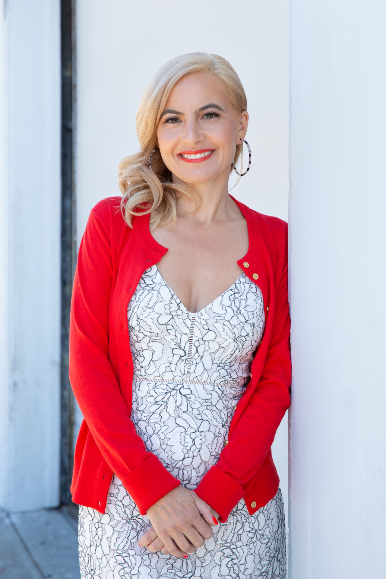 blonde-red-sweater-professional April Hirschman in a white dress with a red sweater leaning against a white wall, smiling.